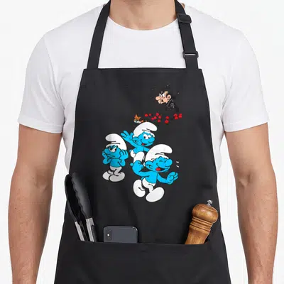 Tablier de cuisine noir avec trois Schtroumpfs souriants courant devant un buisson où se cache Gargamel.