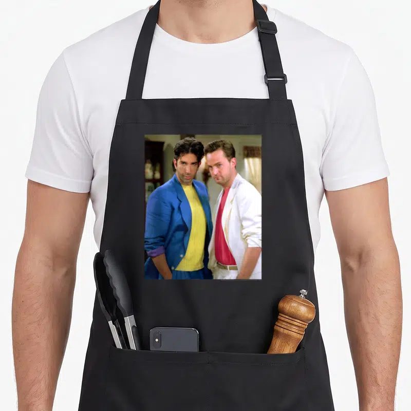 Tablier de cuisine noir avec une illustration colorée de deux hommes en vestes des années 80 posant côte à côte.