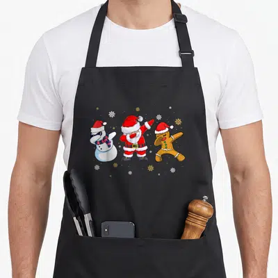 Tablier de cuisine noir avec Père Noël, bonhomme de neige et biscuit faisant le dab sous des flocons.