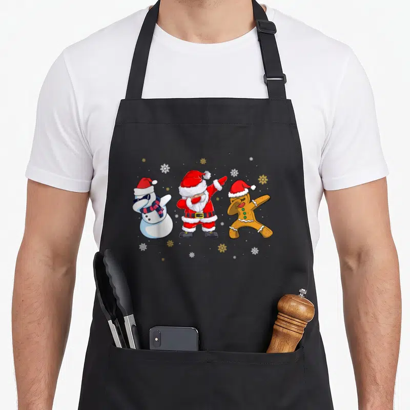 Tablier de cuisine noir avec Père Noël, bonhomme de neige et biscuit faisant le dab sous des flocons.
