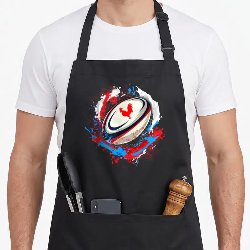 Tablier de cuisine noir avec un ballon de rugby blanc portant un coq rouge et des éclaboussures tricolores.