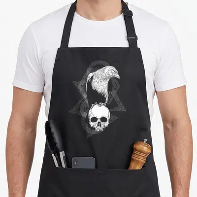 Tablier de cuisine noir avec illustration blanche d'un corbeau posé sur un crâne viking entouré de motifs géométriques linéaires.