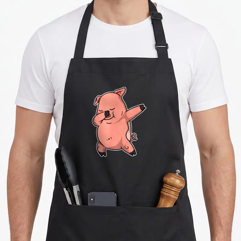 Tablier de cuisine noir avec illustration d'un cochon rose effectuant une pose de dab stylisée sur fond uni.
