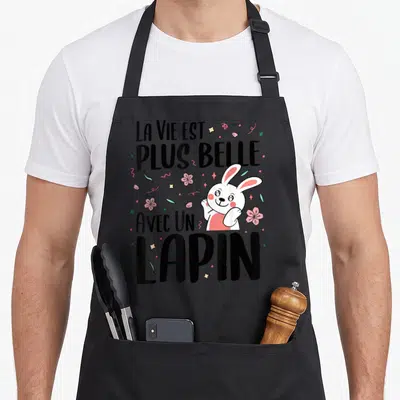 Tablier de cuisine noir avec un lapin blanc souriant, des fleurs roses et le texte La Vie Est Plus Belle Avec Un Lapin en noir.