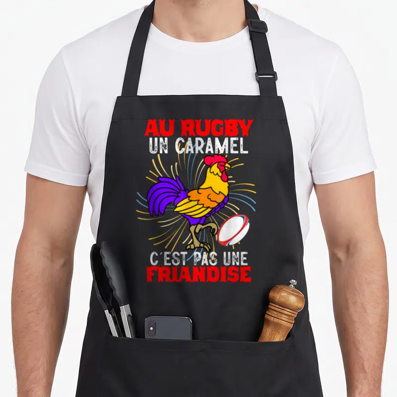 Tablier de cuisine noir avec un coq coloré jouant au rugby et le texte Au Rugby Un Caramel C'est Pas Une Friandise imprimé en rouge et blanc.