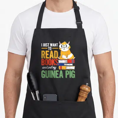 Tablier de cuisine noir avec un cochon d'Inde orange portant des lunettes posé sur une pile de livres colorés.