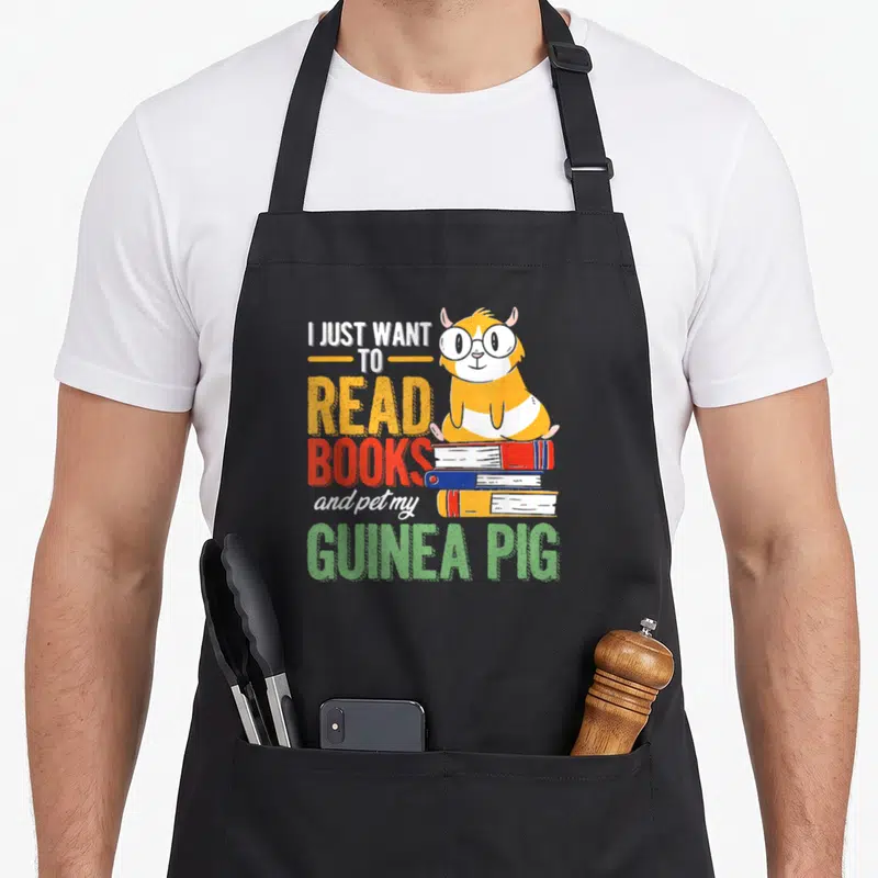 Tablier de cuisine noir avec un cochon d'Inde orange portant des lunettes posé sur une pile de livres colorés.