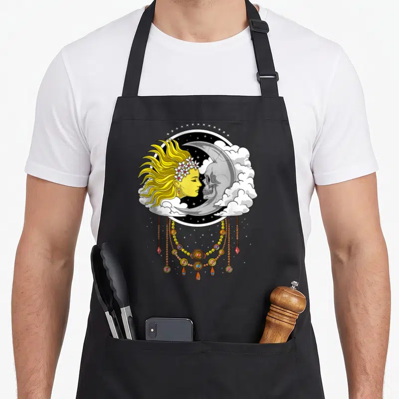 Tablier de cuisine noir avec illustration d'un crâne, d'une lune et d'un soleil entourés de nuages et de bijoux.