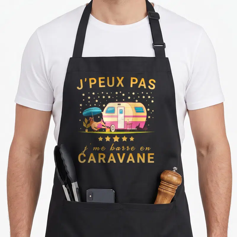Tablier de cuisine noir avec illustration d'un canard en chaise longue devant une caravane rose et texte jaune doré.