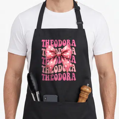 Tablier de cuisine noir avec grand nœud rose à pois blancs et le mot Theodora répété en arrière-plan.