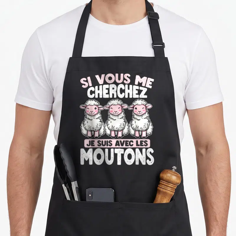 Tablier de cuisine noir avec trois moutons roses souriants et le texte Si vous me cherchez je suis avec les moutons imprimé en blanc et rose.