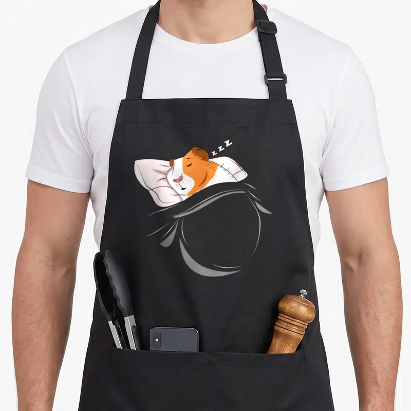 Tablier de cuisine noir avec illustration d'un cochon d'Inde dormeur sous une couverture rose et grise.