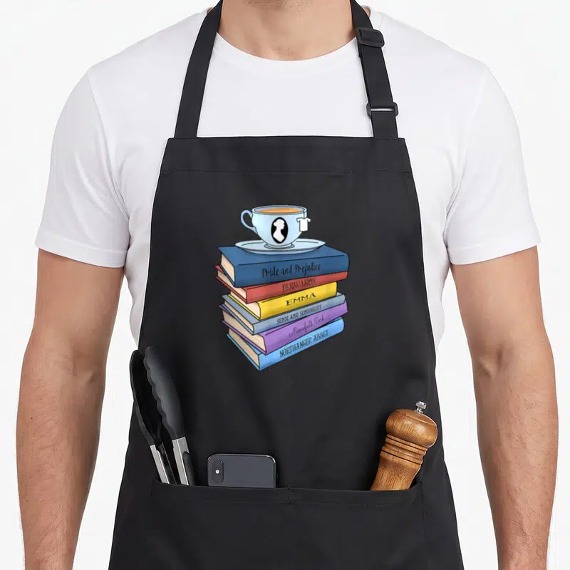 Tablier de cuisine noir avec une illustration d'une pile de livres de Jane Austen surmontée d'une tasse de thé bleue.