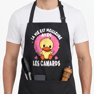 Tablier de cuisine noir avec un canard jaune mignon et l'inscription La vie est meilleure avec les canards sur fond rose.