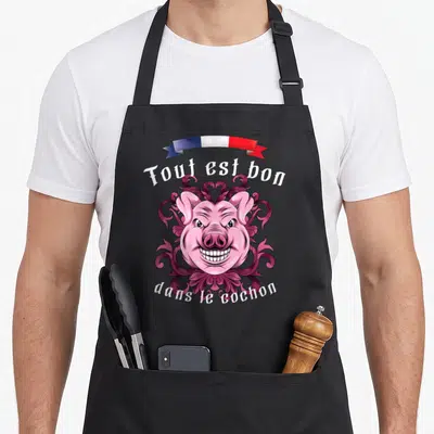Tablier de cuisine noir avec illustration d'un visage de porc rose souriant et texte blanc sur fond gris uni.