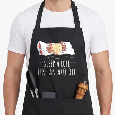 Tablier de cuisine noir avec un axolotl rose dormant sur un oreiller et du texte humoristique en anglais.