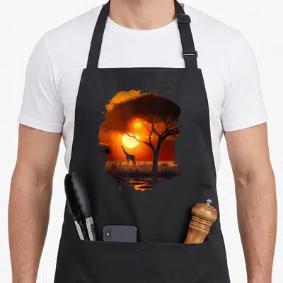 Tablier de cuisine noir avec illustration d'une girafe silhouette sous un arbre au coucher de soleil orange vif.