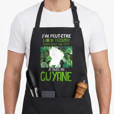 Tablier de cuisine noir avec silhouette de la Guyane entourée de plantes tropicales vertes et texte humoristique blanc et vert.