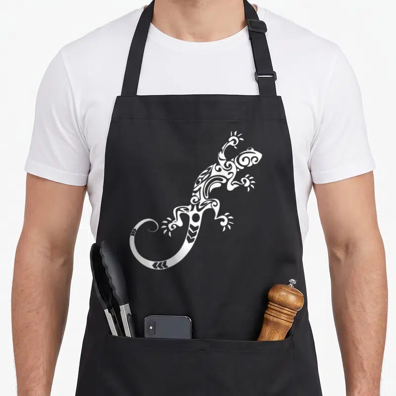 Tablier de cuisine noir avec une illustration blanche d'un lézard au motif tribal et des volutes décoratives sur le corps.