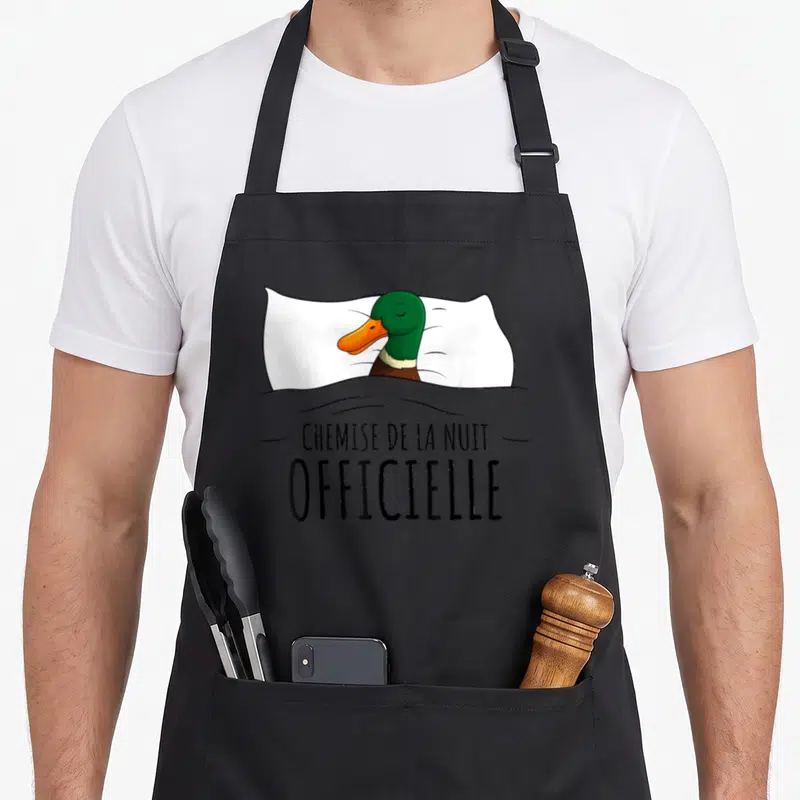 Tablier de cuisine noir avec illustration d'un canard vert dormant sous une couette grise sur un oreiller blanc.