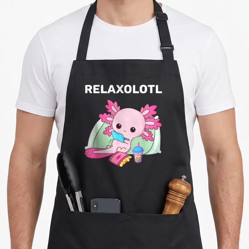 Tablier de cuisine noir avec un relaxolotl rose assis sur des coussins avec une tablette, des frites et une boisson à la perle.