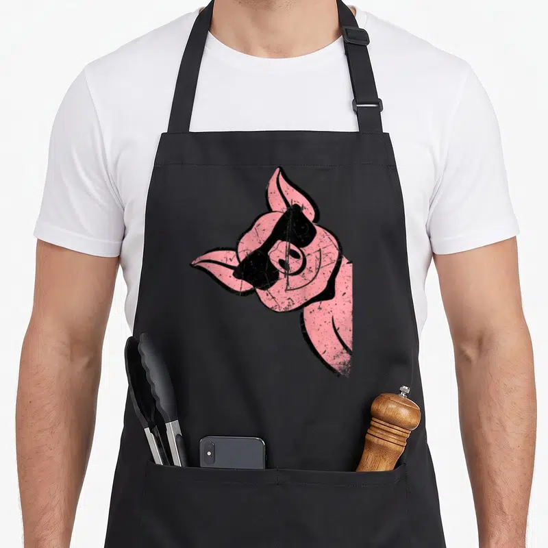 Tablier de cuisine noir avec illustration d'un cochon rose souriant portant des lunettes de soleil noires au style rétro.