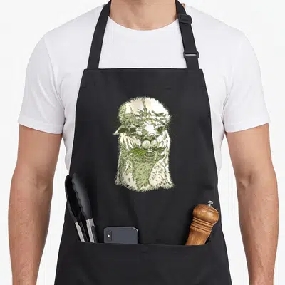 Tablier de cuisine noir avec illustration d'un alpaga au visage dessiné à la main et une feuille verte dans la bouche.