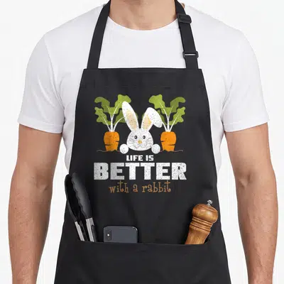 Tablier de cuisine noir avec illustration d'un lapin blanc souriant entre deux carottes orange et le texte Life is Better with a rabbit.