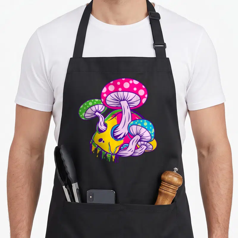 Tablier de cuisine noir avec crâne jaune schmelzen et champignons colorés roses verts et bleus sur fond uni.