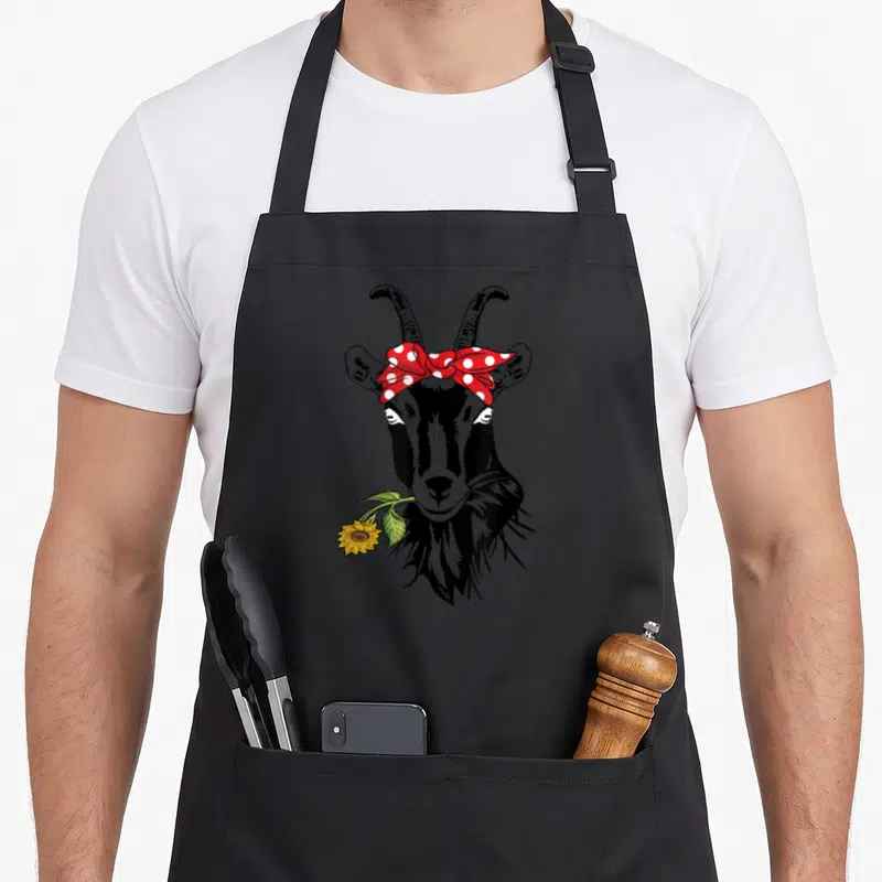 Tablier de cuisine noir avec tête de chèvre au bandana rouge à pois et une fleur de tournesol jaune dans la bouche.