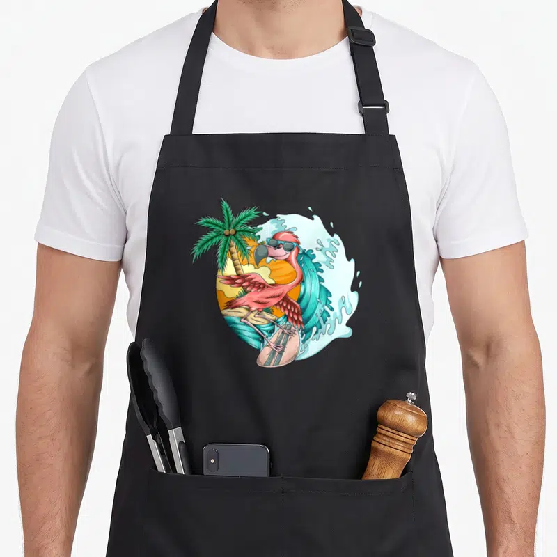 Tablier de cuisine noir avec un flamant rose surfeur portant des lunettes de soleil devant une vague et un palmier.