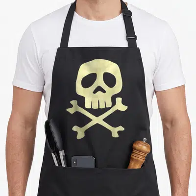 Tablier de cuisine noir avec un crâne et des os croisés imprimés en beige texturé au centre du vêtement.