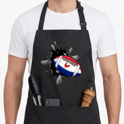 Tablier de cuisine noir avec un ballon de rugby tridimensionnel percant le fond, arborant un coq rouge sur les bandes bleue, blanche et rouge.