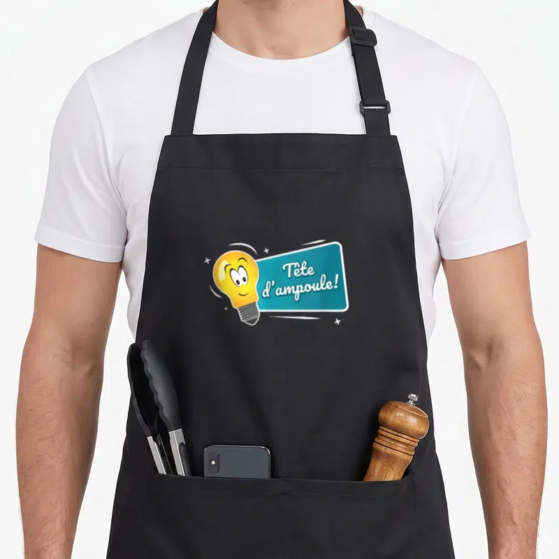 Tablier de cuisine noir avec une ampoule jaune souriante et une étiquette bleue portant l'inscription Tête d'ampoule.
