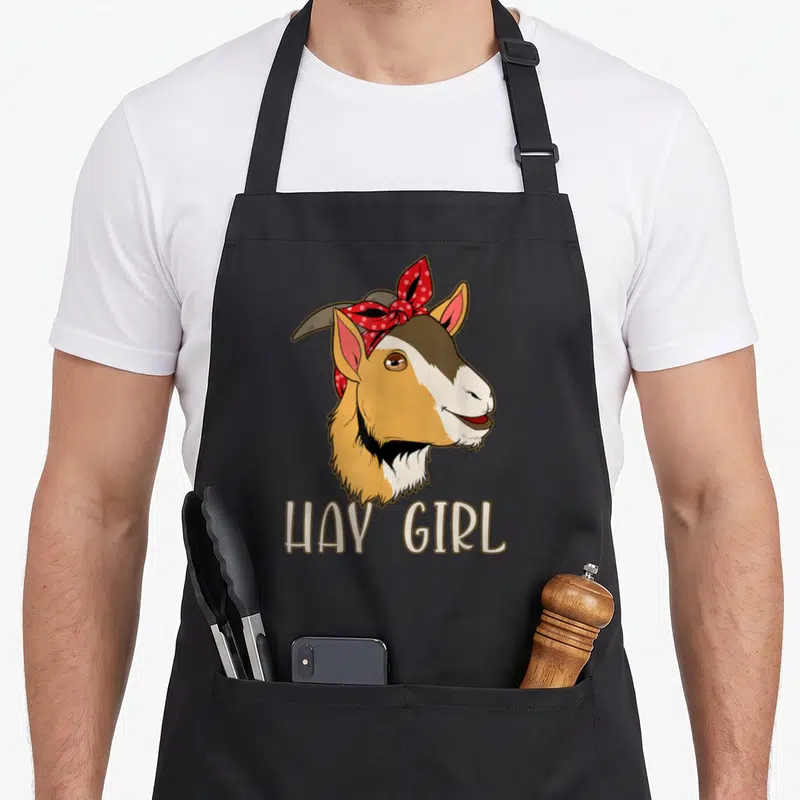Tablier de cuisine noir avec tête de chèvre souriante et foulard à pois rouges, texte HAY GIRR en bas.