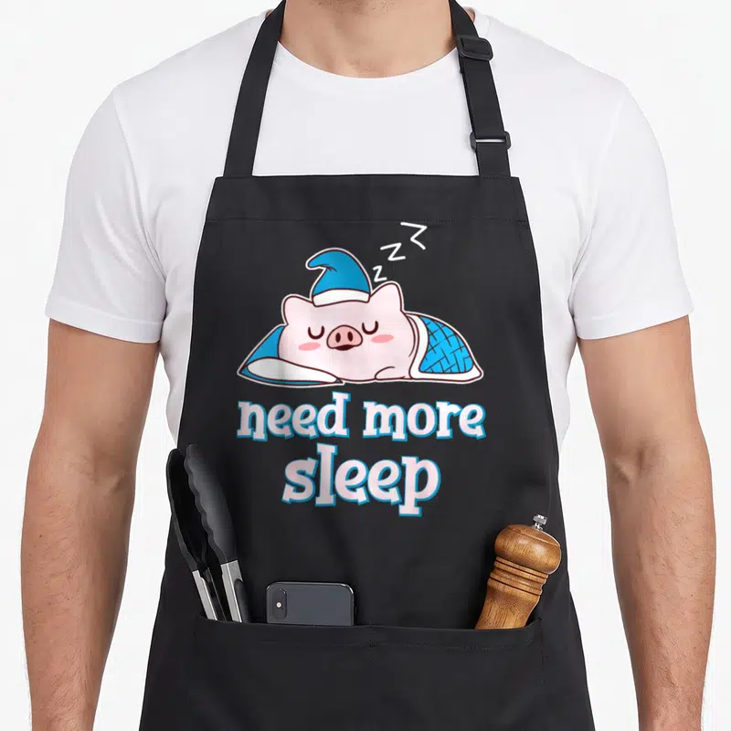 Tablier de cuisine noir avec un cochon rose dormant portant une bonnet bleu et le texte need more sleep en dessous.