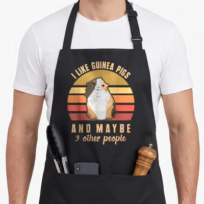 Tablier de cuisine noir avec illustration rétro d'un cochon d'Inde tricolore sur fond de soleil couchant aux bandes orange et rouge.