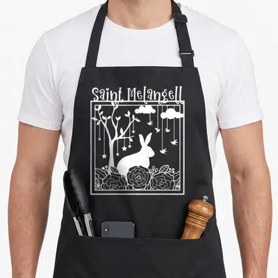 Tablier de cuisine noir avec illustration blanche d'un lapin sous un arbre orné d'étoiles et de fleurs.