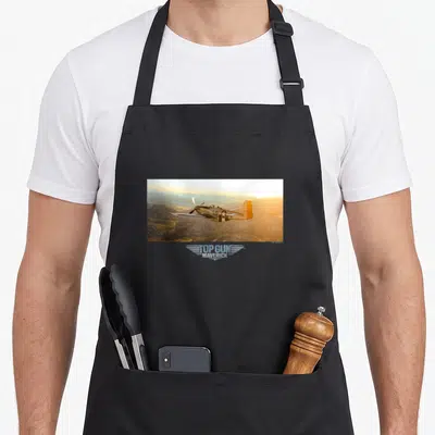 Tablier de cuisine noir avec un avion de chasse P-51 Mustang volant sur des montagnes au coucher de soleil et le logo Top Gun Maverick.