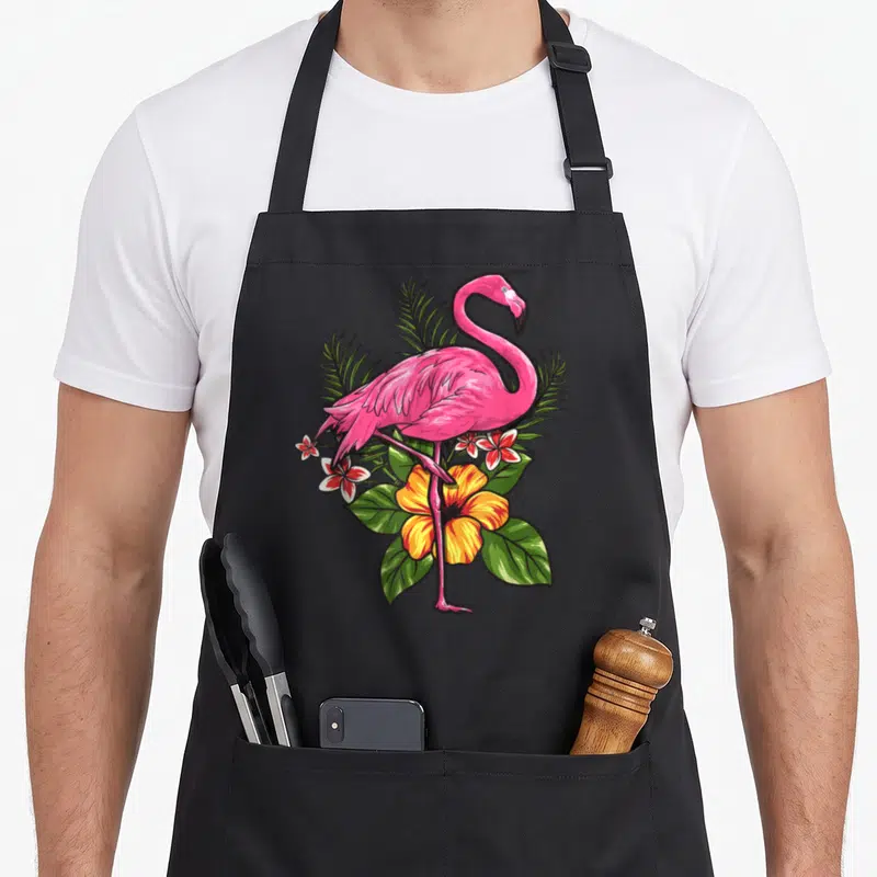 Tablier de cuisine noir avec un flamant rose rose vif entouré de fleurs tropicales et de feuilles de palmier.