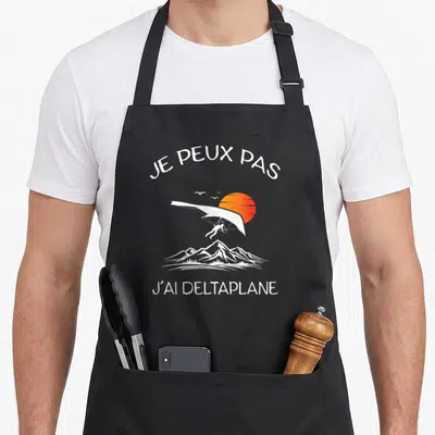 Tablier de cuisine noir avec illustration de deltaplane, soleil orange et montagnes blanches sous le texte Je peux pas j'ai deltaplane.