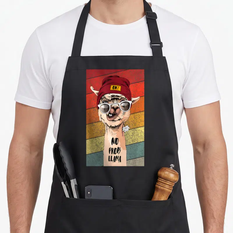 Tablier de cuisine noir avec illustration d'un lama portant des lunettes blanches et un bonnet rouge sur fond coloré.