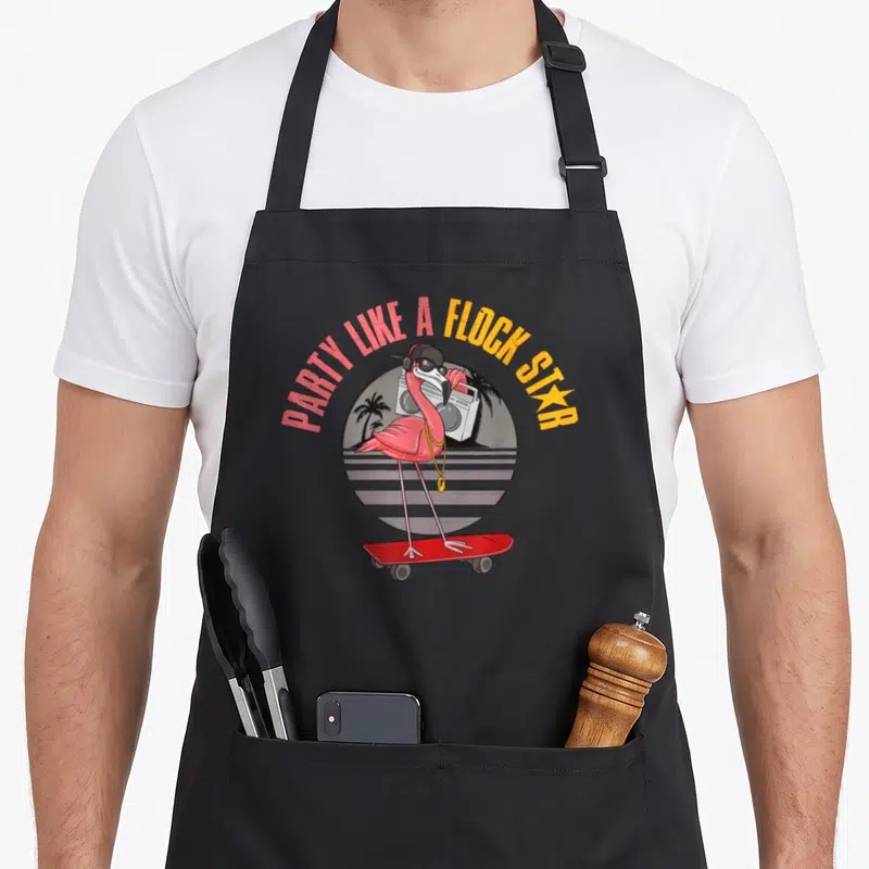Tablier de cuisine noir avec un flamant rose rose vif sur skateboard rouge portant des écouteurs et une chaîne dorée devant une boombox.