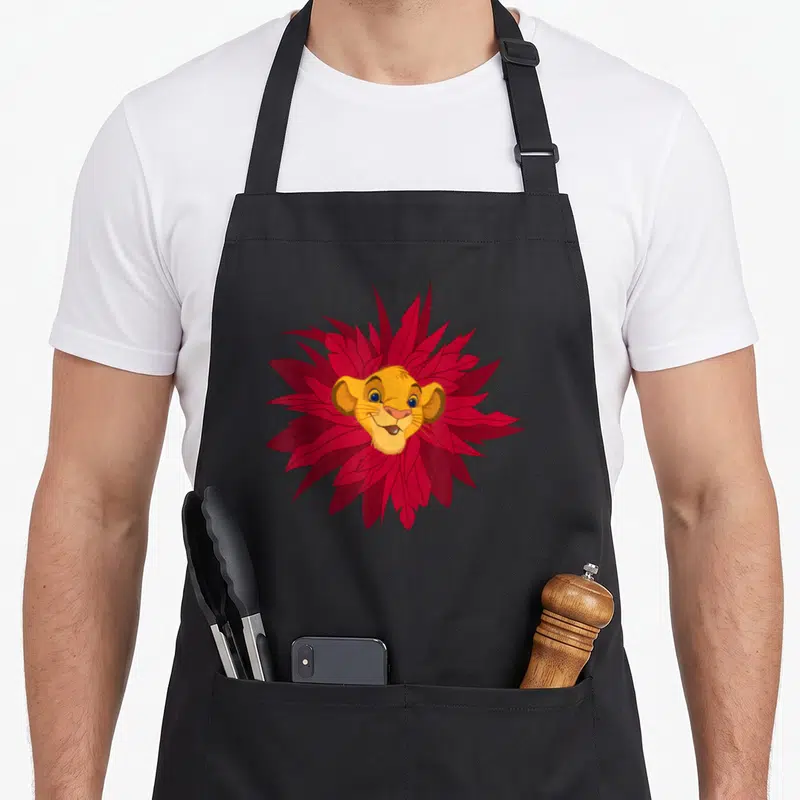 Tablier de cuisine noir avec le visage de Simba entouré de feuilles rouges stylisées formant une mane florale.