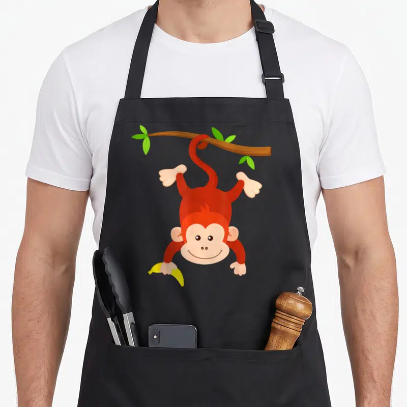 Tablier de cuisine noir avec un singe rouge accroché à une branche tenant une banane jaune.