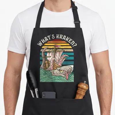Tablier de cuisine noir avec illustration du kraken attaquant un voilier sous le texte What's Kraken sur fond de soleil rayé.
