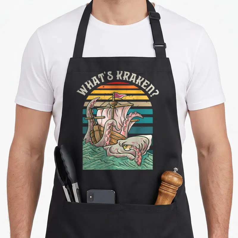Tablier de cuisine noir avec illustration du kraken attaquant un voilier sous le texte What's Kraken sur fond de soleil rayé.