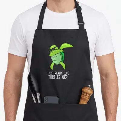 Tablier de cuisine noir avec une tortue verte aux lunettes noires et un texte humoristique en anglais.