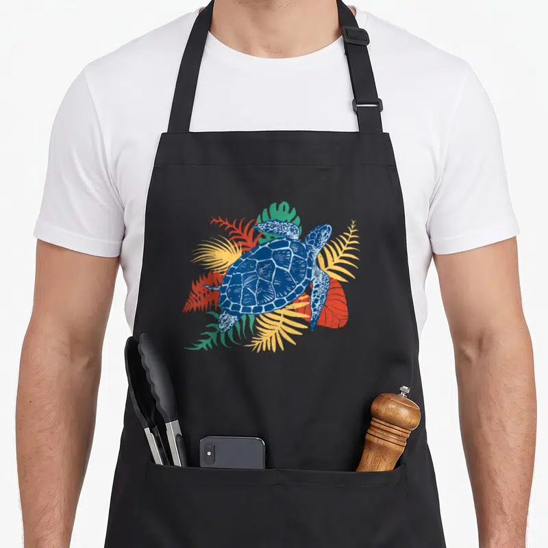 Tablier de cuisine noir avec illustration d'une tortue de mer bleue et de feuilles tropicales multicolores.