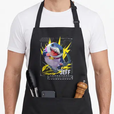 Tablier de cuisine noir avec illustration colorée de Jeff le requin terrestre aux éclairs jaunes et inscription THE LAND SHARK en bas.